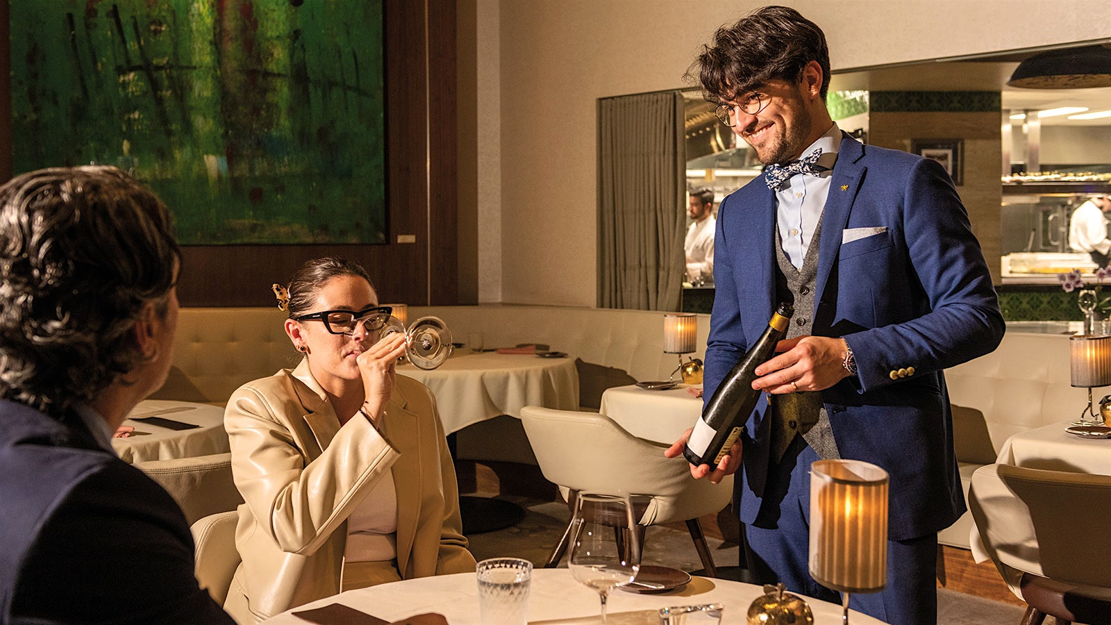 Gabriel Kreuther wine director Aukai Bell displaying a bottle of wine for two patrons in the modern-design dining room of Gabriel Kreuther, with a view into the kitchen, as one of the patrons sips the wine